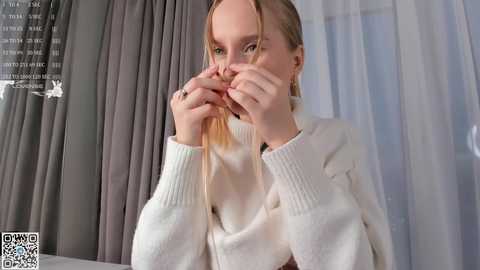 Media: Video of a young woman with blonde hair, wearing a white cardigan, playfully holding her hair, in a brightly lit room with grey curtains and white sheer drapes.