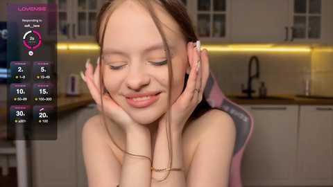 Media: A young woman with fair skin, brown hair, and closed eyes, smiling, in a modern kitchen with white cabinets, a sink, and a pink gaming chair.