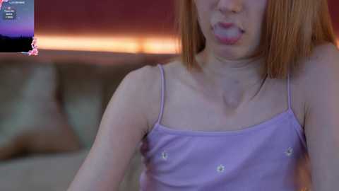 Media: A video of a slim, light-skinned woman with short, straight, reddish-brown hair. She wears a lavender spaghetti-strap top with small buttons. She's licking her lips, and the background shows a blurred, warm-toned setting.