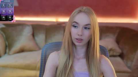 Media: Video of a young, slender Caucasian woman with long blonde hair, wearing a light purple top, sitting on a grey chair in a dimly lit room with a pinkish wall and beige couch.