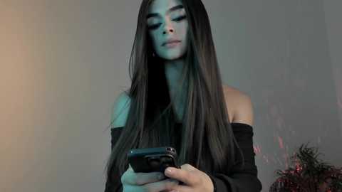 Media: Video of a woman with long, straight black hair, wearing an off-shoulder black top, looking down at a smartphone. Background features a blurred, muted color palette.