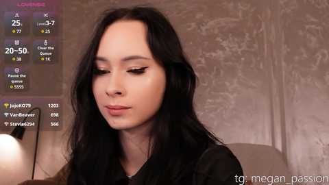 Media: Video of a young woman with long black hair, wearing black clothing, looking downward, in a dimly lit room with a textured beige wall. A digital overlay displays social media statistics.