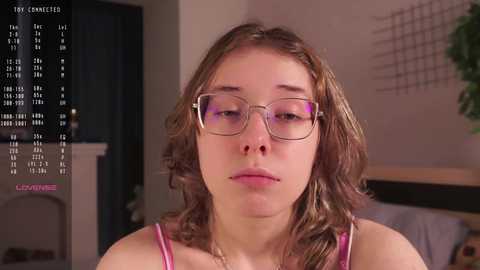 Media: Video of a young Caucasian woman with wavy brown hair, wearing glasses, and a pink tank top, sitting indoors with a calendar overlay in the background.