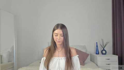 Media: Video of a young woman with long brown hair, wearing a white off-shoulder blouse, standing in a minimalist bedroom with white walls, a bed, and a blue vase.