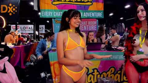 Media: Video of a young woman in a yellow bikini, smiling, at a colorful convention booth with various banners, people, and costumes in the background.