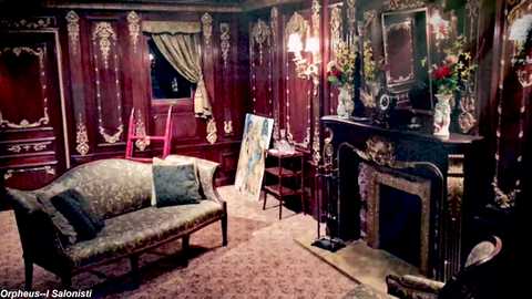 Media: Video of a lavish Victorian-style living room with dark wood paneling, ornate fireplace, floral-patterned sofa, red chairs, and a vintage painting on easel, adorned with rich drapes and elegant floral arrangements.