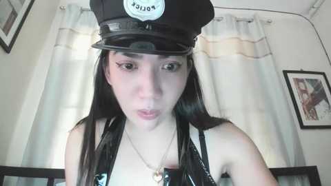 Media: A video of a young woman with long black hair, wearing a black police hat and a black leather jacket, standing in front of a window with white curtains and framed pictures on the walls.