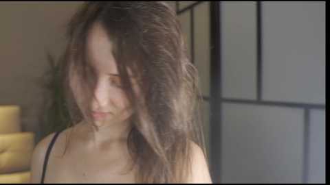 Media: Video of a young woman with long, messy brown hair, wearing a black tank top, standing indoors next to a yellow couch and shoji screen.