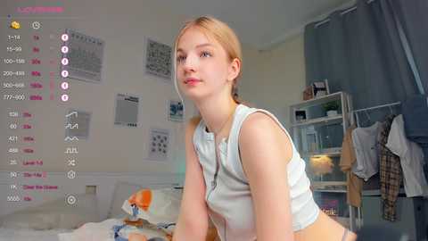 Media: Video of a young woman with light skin and blonde hair, wearing a white crop top, sitting on a bed in a cluttered bedroom.