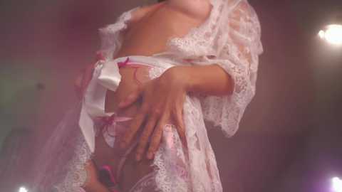 Media: Video of a light-skinned woman wearing a sheer, white lace robe with pink satin bows, revealing her small breasts and pink panties. She poses suggestively, with her left hand on her hip and right hand near her crotch. The background is blurred, with soft lighting creating a sensual atmosphere.