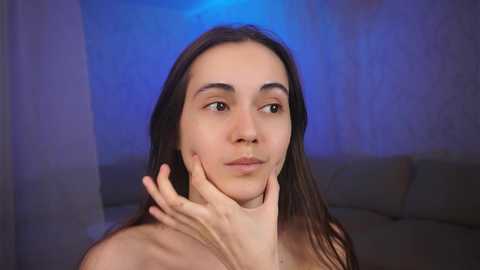 Media: Video of a topless young woman with fair skin, long brown hair, and a neutral expression, touching her face. Background features a soft blue light and a gray sofa.