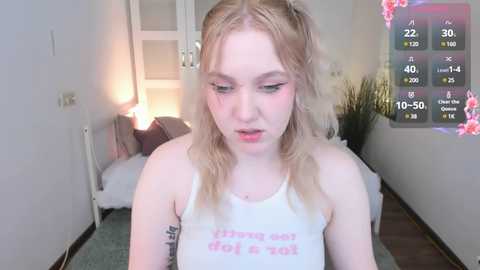 Media: A video of a young, fair-skinned, blonde-haired woman with a tattoo on her arm, wearing a white tank top with the phrase \"Thirsty as fuck\" in pink. She stands in a dimly lit bedroom with a green carpet, a window, and a digital clock displaying 22:50.