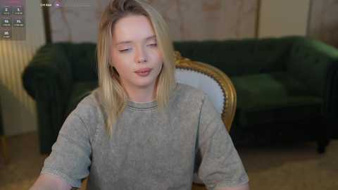 Media: Video of a young blonde woman with fair skin, wearing a gray sweater, sitting indoors on a green sofa with a wooden armchair in the background.