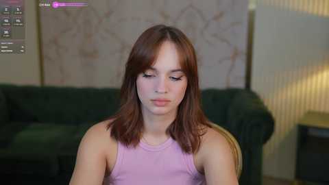 Media: A video of a young woman with shoulder-length brown hair, wearing a light purple tank top, sitting indoors with a green couch and beige walls in the background.