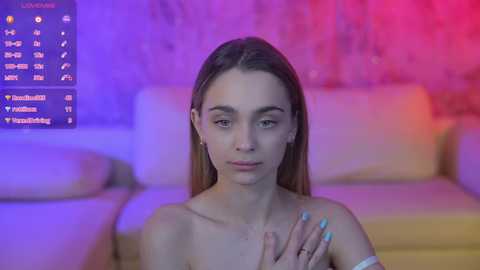 Media: Video of a topless, fair-skinned woman with long brown hair, wearing a silver bracelet, standing in a dimly lit room with purple and pink lighting, against a blurred background of a white sofa.