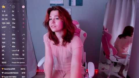 Media: Video of a young woman with red hair and fair skin, wearing a sheer pink top, sitting in a pink gaming chair, in a dimly lit, cozy bedroom.