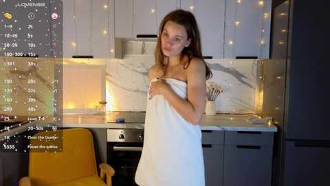 Media: A video of a young woman with fair skin, long brown hair, and a slender physique, wrapped in a white towel, standing in a modern kitchen with white cabinets and warm lighting.