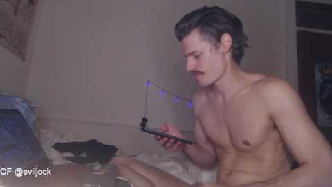 Media: Video of a shirtless, slender young man with curly brown hair, wearing a necklace, holding a phone, sitting on a messy bed with blue string lights, in a dimly lit room.