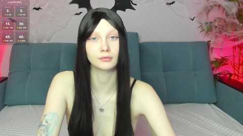 Media: Video of a pale, thin woman with long black hair, wearing a black headband with bat wings, sitting on a bed with blue cushions and a white sheet. Background includes a wall with bat stickers and a plant.