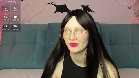 Media: Video of a pale-skinned woman with long black hair and red lipstick, wearing black cat ears, glasses, and a black sleeveless top. She's seated on a blue couch with a pinkish wall in the background.