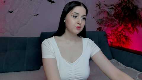 Media: A video of a young woman with long black hair, wearing a white top, sitting on a dark sofa in a room with a floral wall mural and a red-lit corner.