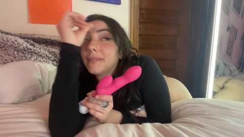 Media: A video of a woman lying on a bed, wearing a black long-sleeve top, holding a pink vibrator, with a bedspread, wall art, and wooden door in the background.