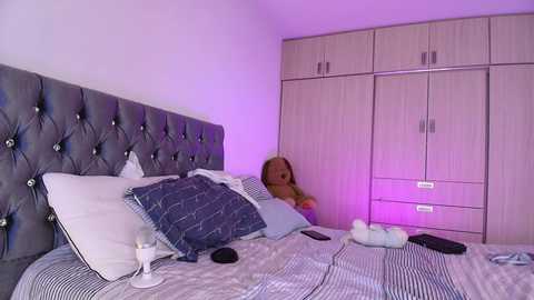 Media: A video of a modern bedroom with a woman in a blue dress lying on a bed with white pillows and a striped blanket. A large, tufted grey headboard is behind her. The room has a purple lighting ambiance.