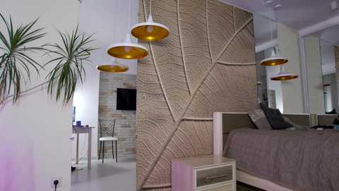 Media: Modern bedroom with beige leaf-patterned wall, white bed, gray bedding, two white chairs, desk, potted plant, hanging lamps, and minimalist decor.