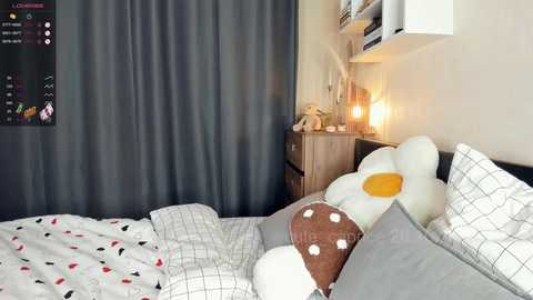 Media: A video of a cozy, modern bedroom featuring a white bed with gray checkered sheets, a plush white and yellow pillow, and a brown pillow with white polka dots. A gray curtain and a wooden nightstand with a lamp are visible.