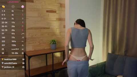 Media: A video of a slender woman with light skin, wearing a blue crop top and white panties, pulling them down, in a modern living room with a wooden wall and beige curtains.