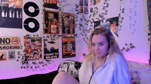 Media: Video of a young woman with blonde hair in a light blue hoodie, seated on a couch with a collage of music posters and album covers in the background.