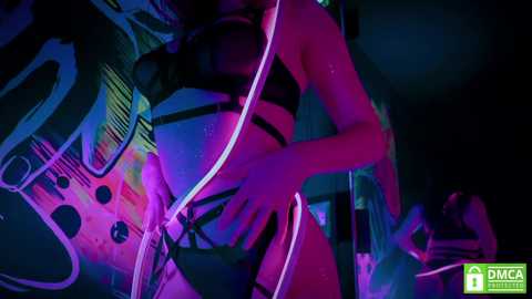 Media: A video of a scantily clad woman with a slender physique, wearing black lingerie, illuminated by neon lights in a dimly lit room with graffiti walls.