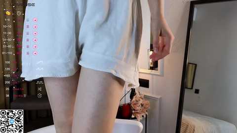 Media: A video of a woman's legs in white shorts, standing in a modern bedroom with a white bed, framed artwork, and a QR code watermark in the bottom left corner.