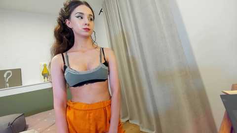 Media: Video of a young woman with long, wavy brown hair wearing a gray bralette and orange high-waisted pants, standing in a modern, minimally decorated bedroom with white walls and a gray curtain.