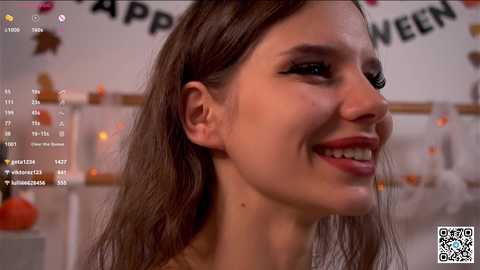 Media: Video of a smiling woman with straight, dark brown hair, fair skin, and light makeup, possibly in her 20s, in a Halloween-themed room with \"Happy Halloween\" banner and festive decorations.