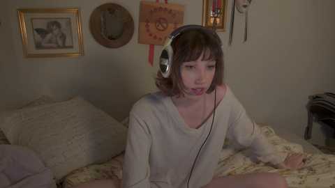 Media: Video of a young woman with short brown hair, wearing a beige sweater and headphones, sitting on a floral-patterned bed in a cozy room with framed artwork on the wall.