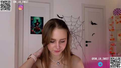 Media: Video of a young, light-skinned woman with long, straight brown hair, wearing a necklace, in a room with Halloween decorations, including a spider web and bats.