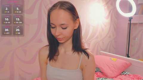 Media: Video of a young, slender, light-skinned Asian girl with straight black hair, wearing a white spaghetti-strap tank top, sitting on a pink bed with a circular lightbulb in the background.