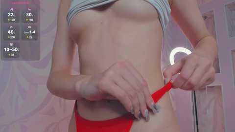 Media: Video of a slender, light-skinned woman lifting her shirt to reveal small breasts and adjusting red panties. Background shows a pink wall and a digital clock display.