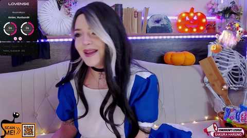 Media: Video of a woman with long, black and white hair, wearing a blue and white Alice in Wonderland costume, smiling while seated on a white couch in a cozy room with festive decor.