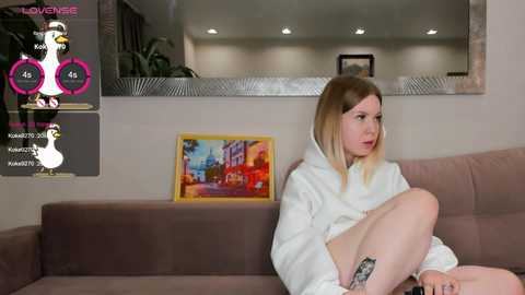 Media: A video of a blonde woman in a white bathrobe, sitting on a beige couch in a modern living room, with a colorful painting and a duck logo on the wall.