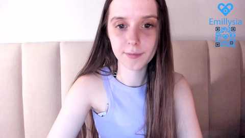 Media: Video of a young woman with long, straight brown hair, fair skin, and a slender physique, wearing a light blue sleeveless top. She is seated against a beige, tufted headboard in a bedroom.