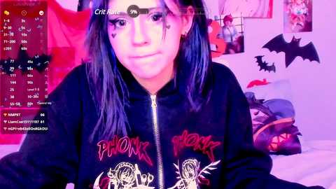 Media: Video of a woman with long, dark hair wearing a black hoodie with metal band logos, sitting in a bedroom with Halloween decorations, including bats and a pumpkin.