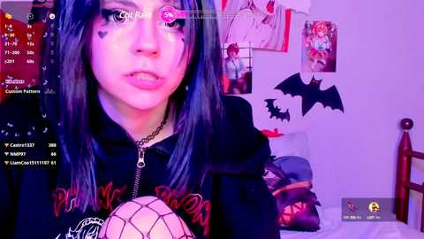 Media: Video of a young woman with long, dark hair, wearing a black hoodie, sitting on a bed. The room is decorated with Halloween-themed decorations, including bats and a framed photo.