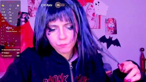 Media: Video of a woman with black hair, wearing a black hoodie with a skull graphic, in a room decorated with anime posters and a bat wall decal. She looks down with a slight smile.