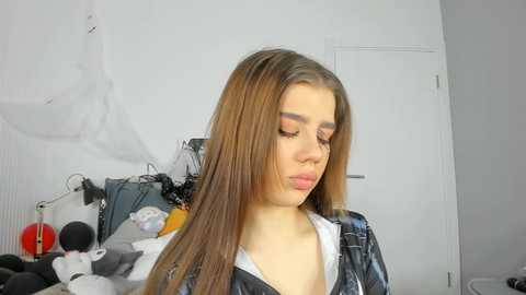 Media: Video of a young woman with straight, light brown hair, wearing a black jacket over a white top, applying makeup in a cluttered bedroom with white walls, a closed door, and a messy desk.