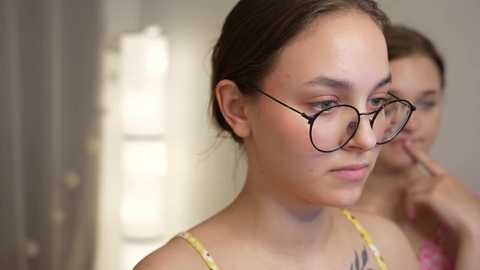 Media: Video of a young Caucasian woman with light skin and brown hair, wearing glasses and a yellow top, standing indoors next to another person.