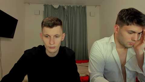 Media: Video of two young men, one in a black shirt, the other shirtless, sitting at a table in a dimly lit room with grey curtains and a black TV screen.