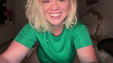 Media: Video of a smiling, light-skinned, blonde woman with shoulder-length hair, wearing a green t-shirt, seated in a dimly lit room with a wooden chair, beige couch, and partially visible window in the background.