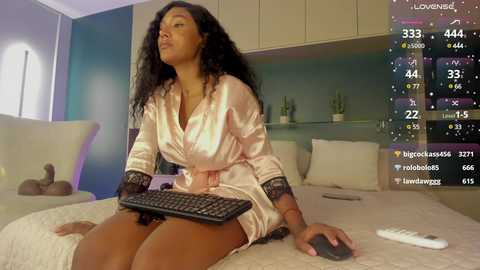 Media: Video of a Black woman with long curly hair in a satin robe, typing on a keyboard in a modern bedroom with white walls and plants.